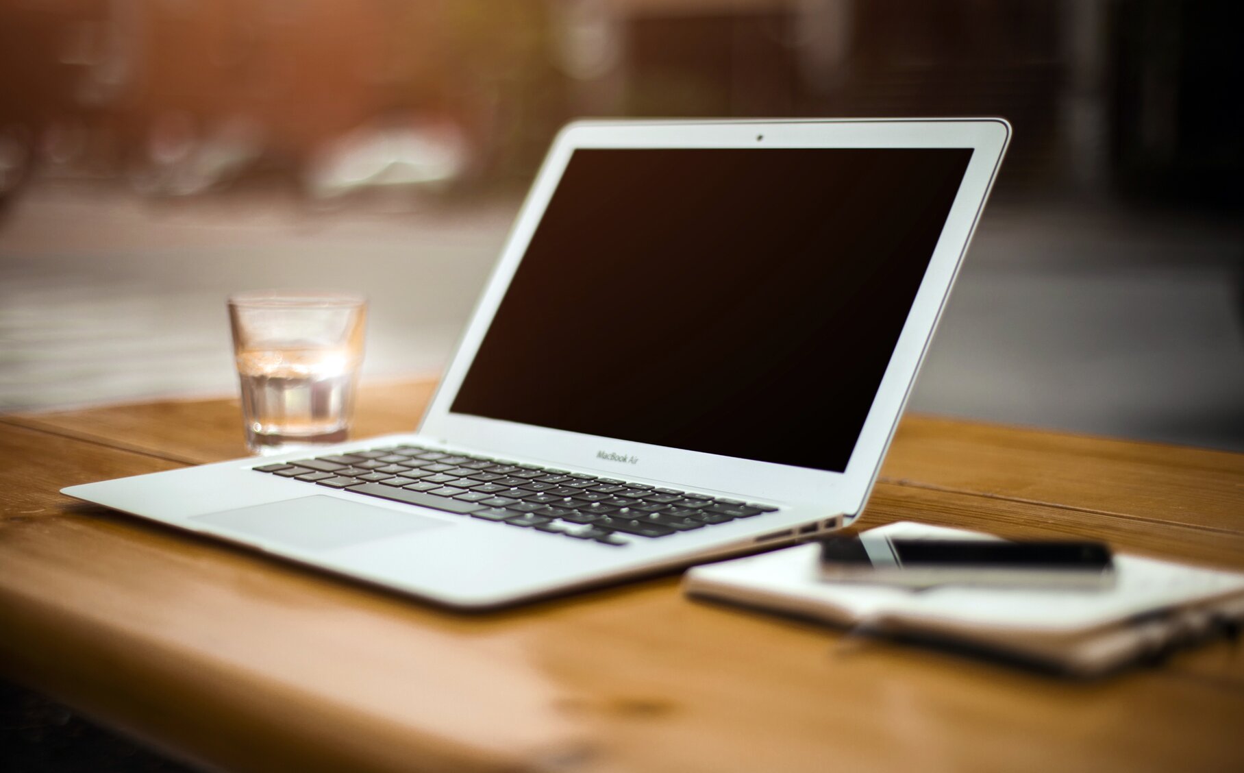 Ein Macbook, ein Glas Wasser, ein Notizblock und ein Handy liegen auf einem Tisch