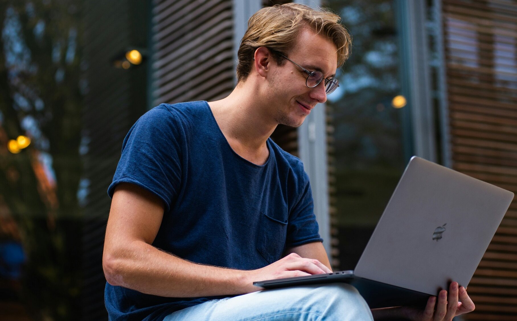 Ein Mann mit Brille sitzt vor einem Holzhaus mit einem aufgeklappten Laptop auf dem Schoß