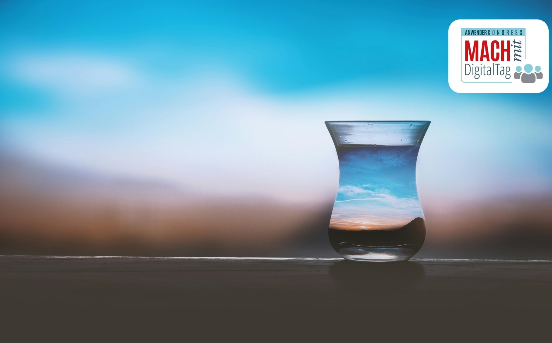 MACHmitDigitalTag 2024 Ein Wasserglas, in dem sich der Sonnenuntergang im unscharfen Hintergrund spiegelt, daneben das Logo vom MACHmitDigitalTag 2024