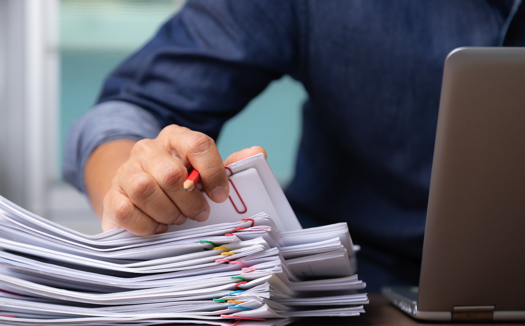 Nahaufnahme einer Person am Laptop, die mit Stift in der Hand einen Papierstapel durchschaut.