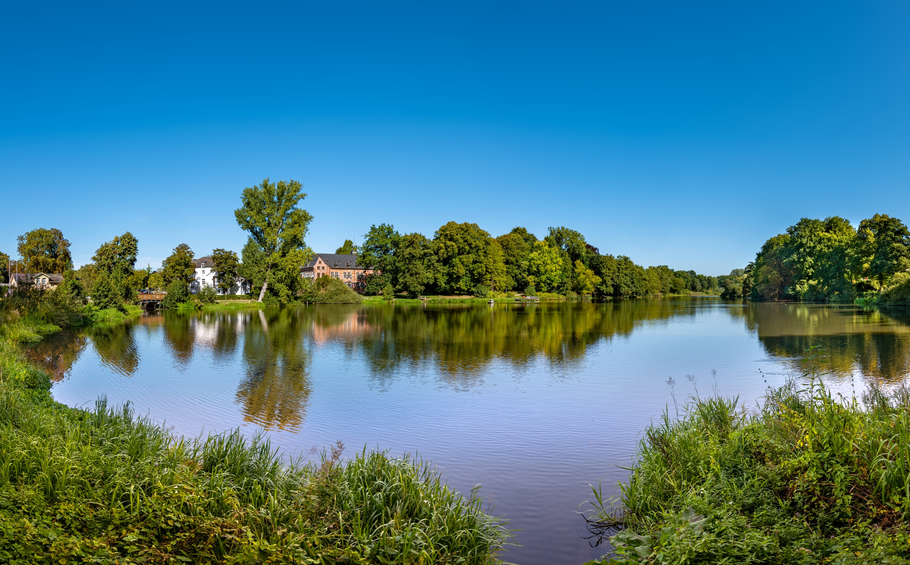 Panoramablick auf Reinbek in Schleswig-Holstein mit Flusslauf, grünen Uferlandschaften, Bäumen und Wohnhäusern unter blauem Himmel. | © foto-select/Shutterstock.com