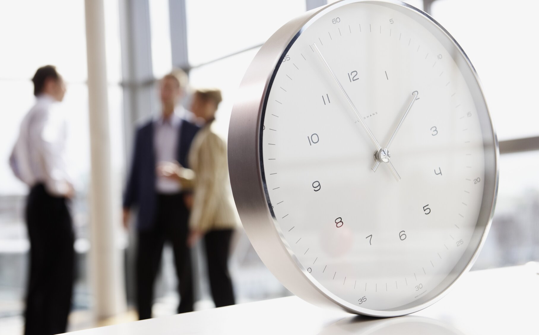 Eine Uhr ist  im Fokus - im Hintergrund verschwommen drei stehende Mitarbeiter.