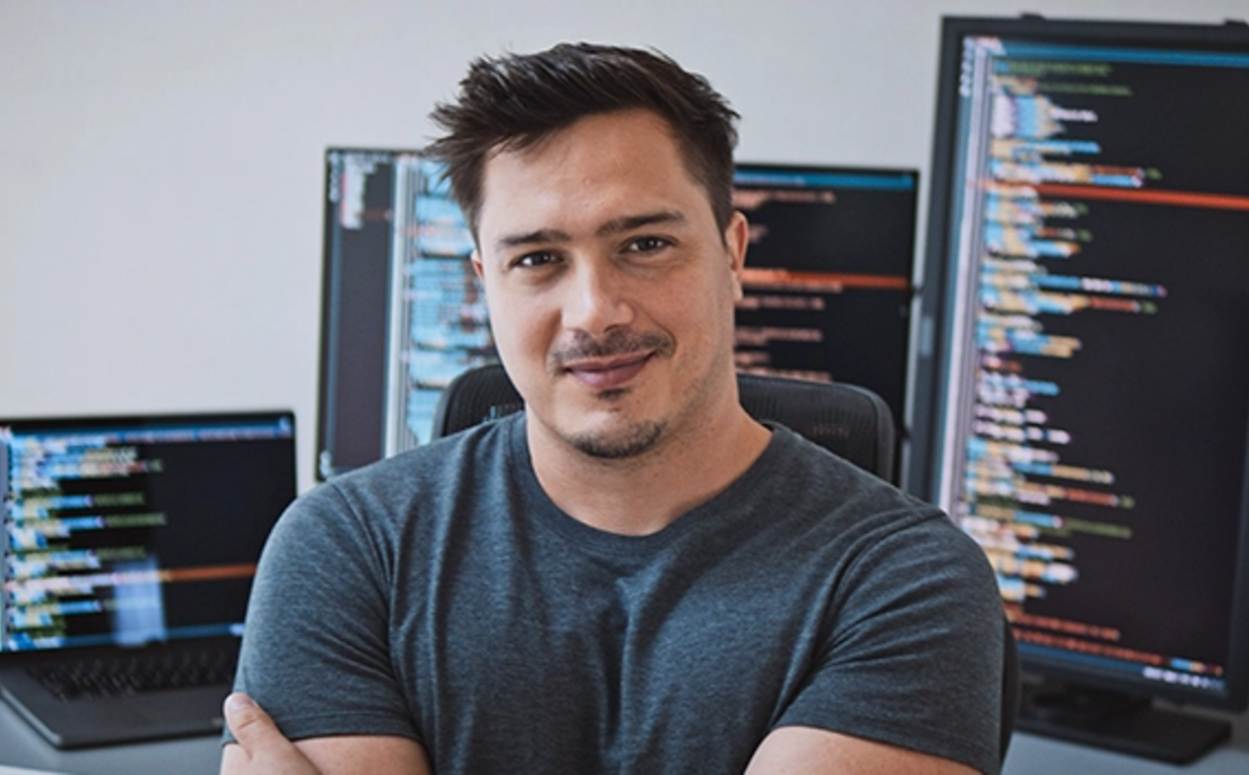 Ein Mann steht mit verschränkten Armen lächelnd vor einem Computerserver inklusive Bildschrim.