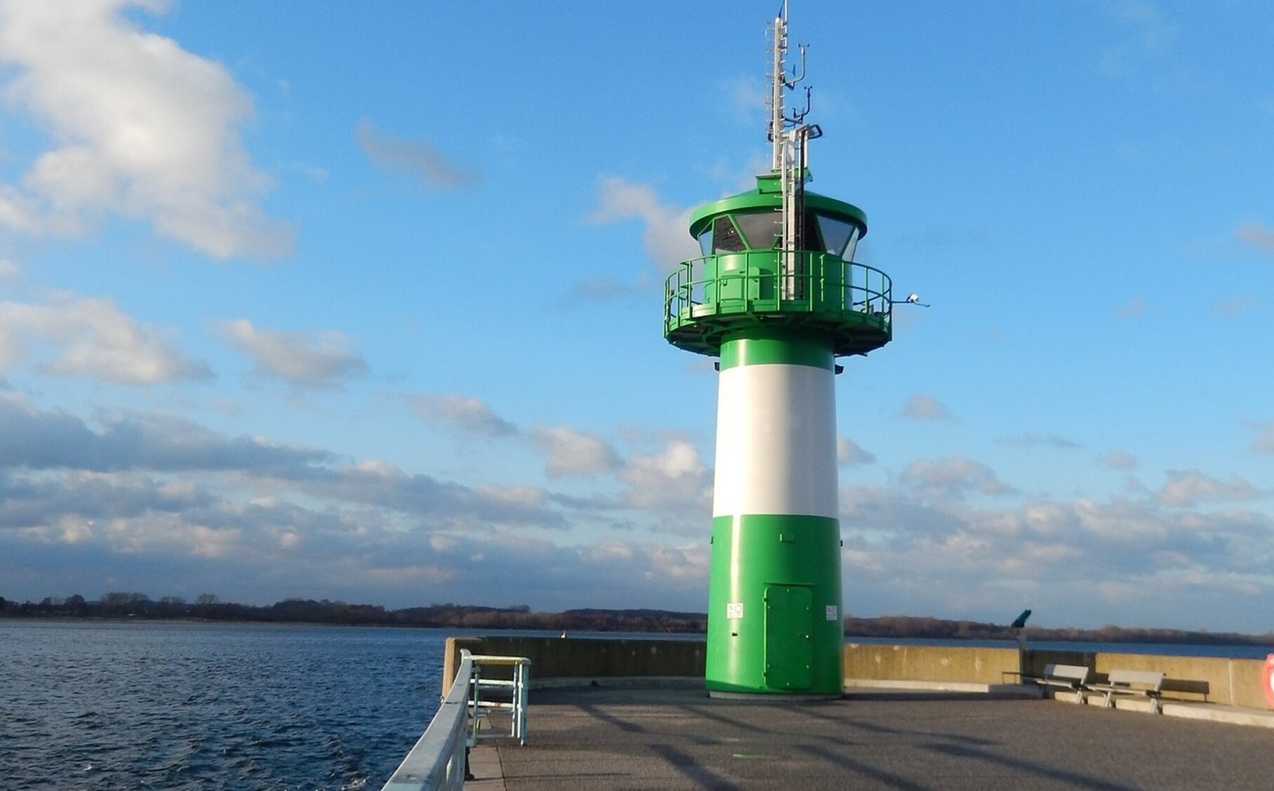 Ein grün-weiß gestreifter Leuchtturm steht am Meer und es ist blauer Himmel mit wenigen Wolken zu sehen.